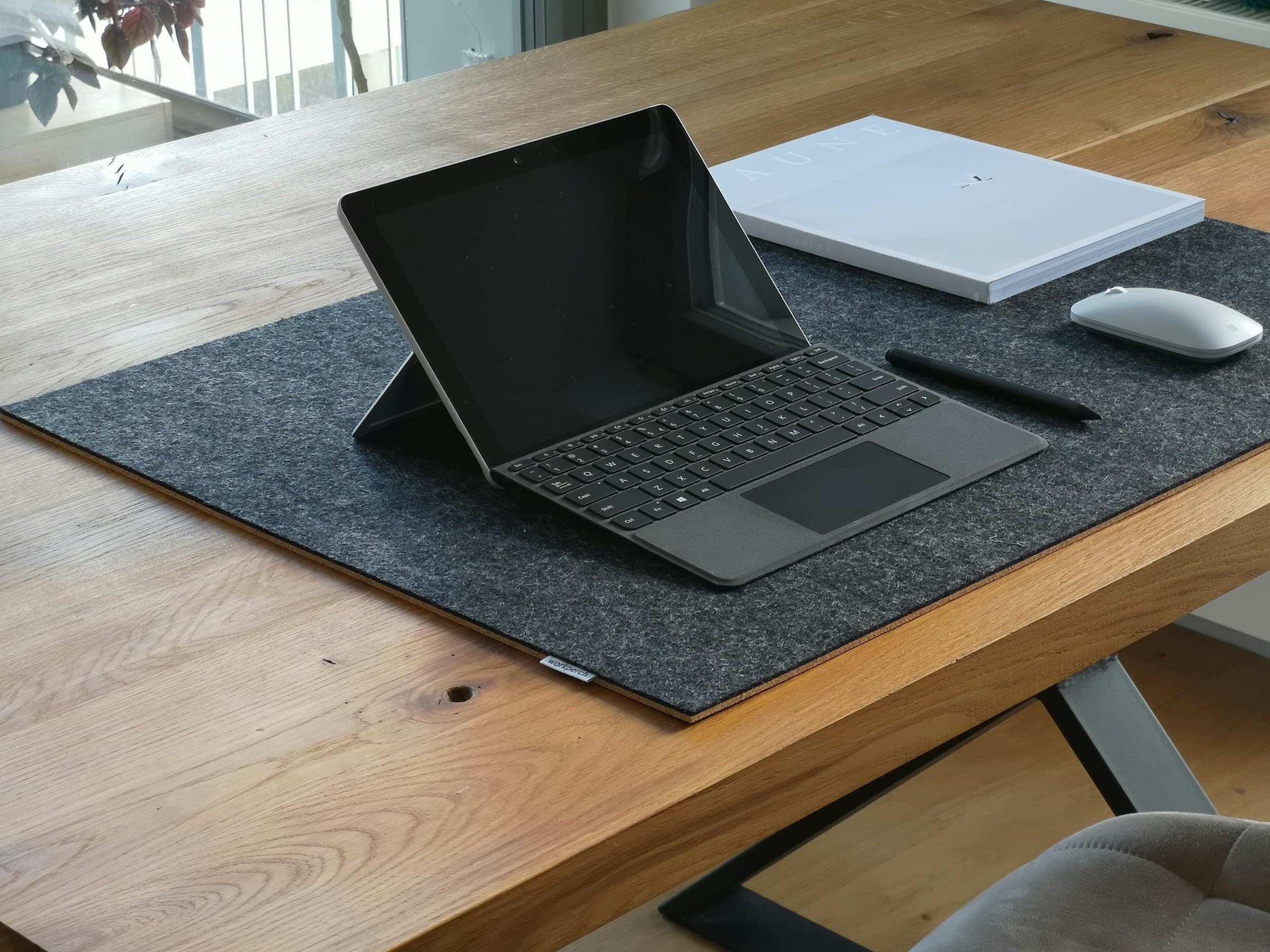 Tablette avec clavier, souris et carnet posés sur un grand tapis de bureau en feutre gris, sur une table en bois