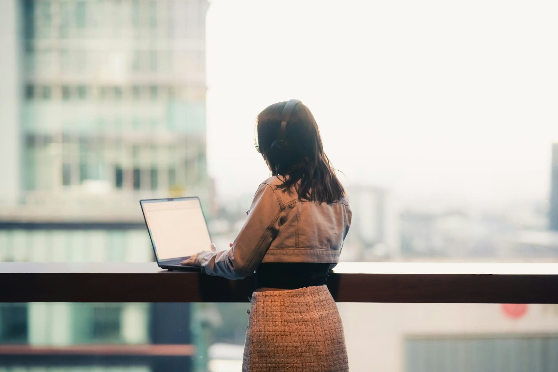 A woman wearing headphones works on a laptop in front of a large window