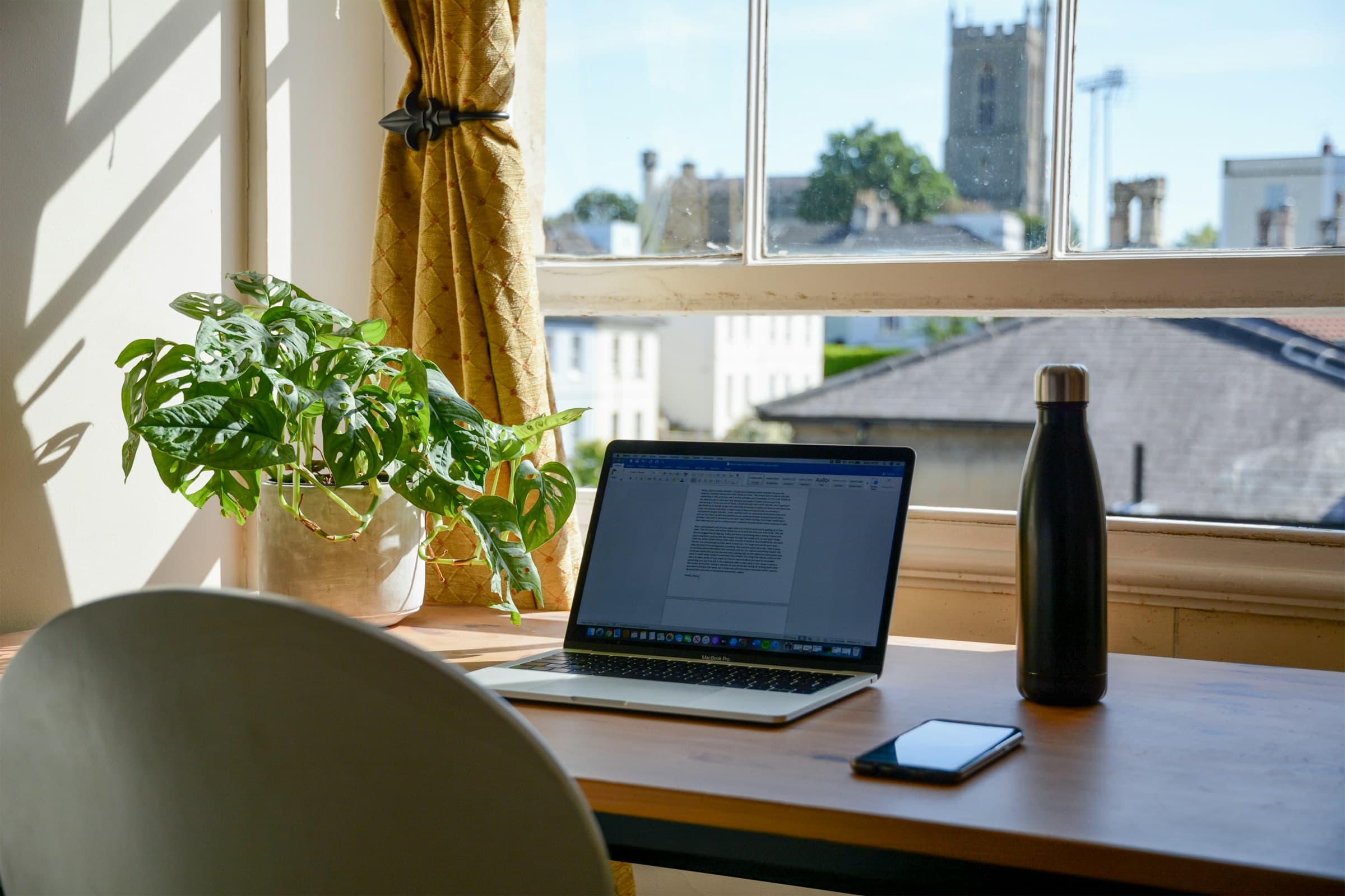 Remote-work desk in summer light with a laptop, curtain, plant, and water bottle by a window