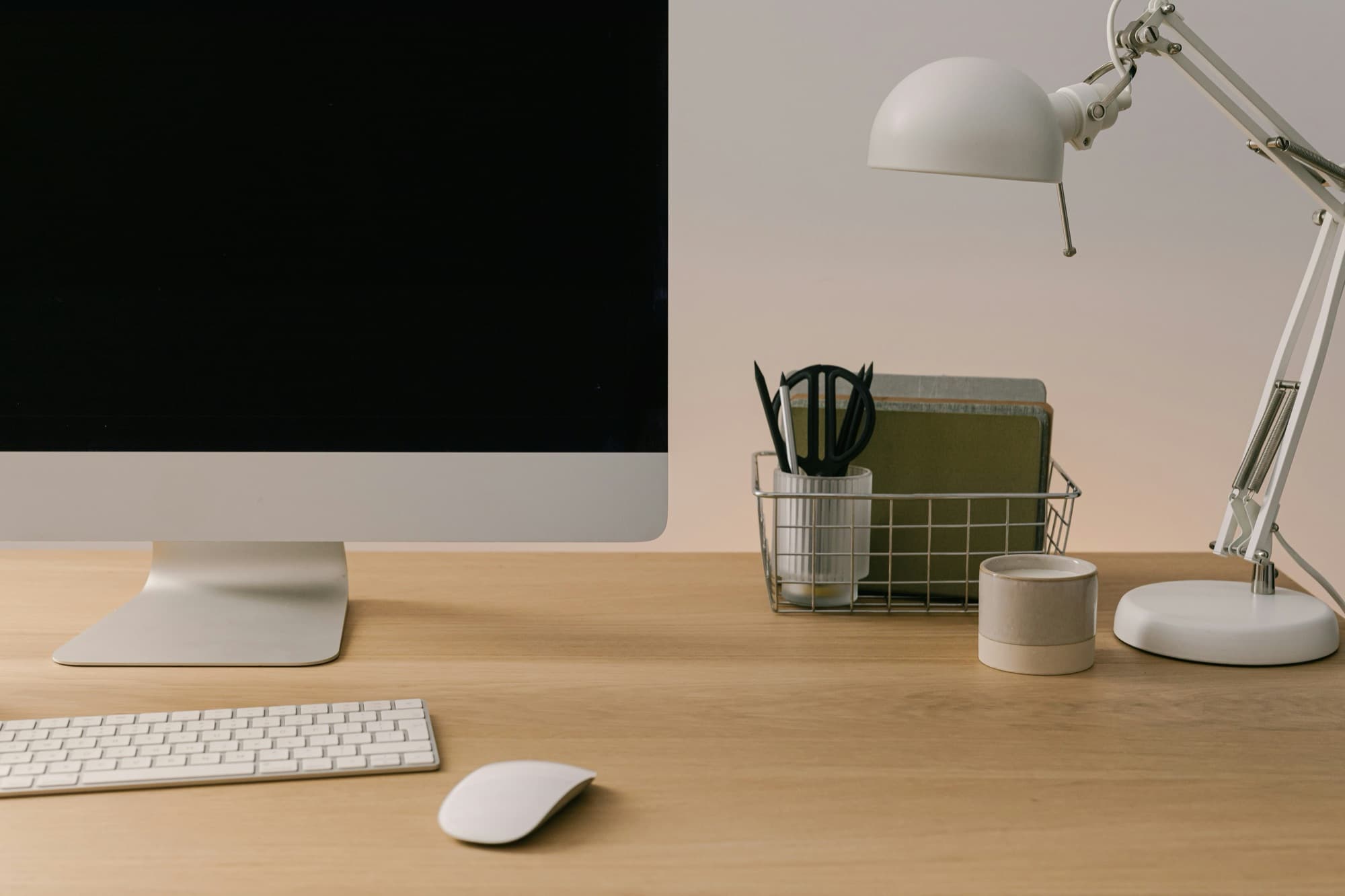Wooden home-office desk with a monitor, keyboard, mouse, white adjustable lamp, and stored stationery
