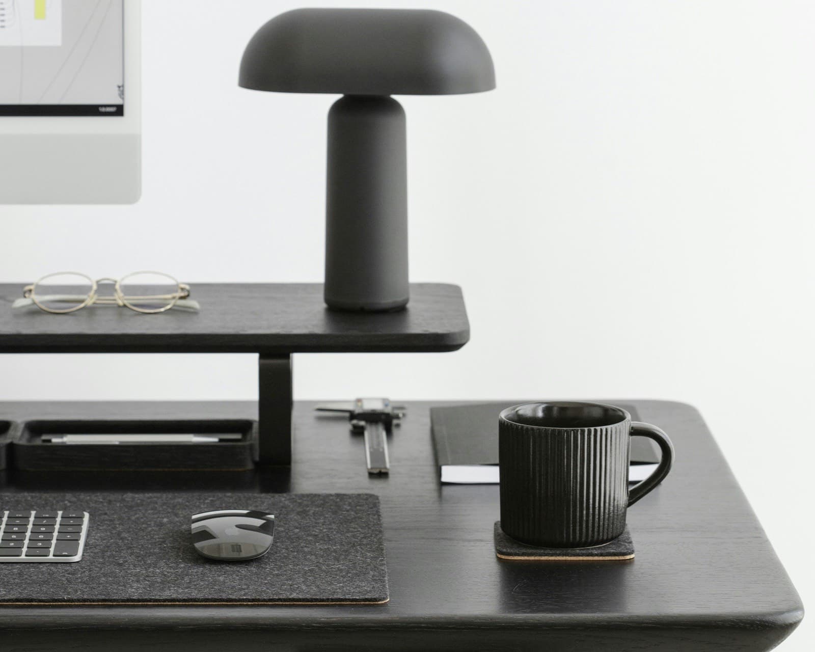 Black desk with a raised shelf holding a lamp and glasses, small items stored underneath, and a mug on the desktop