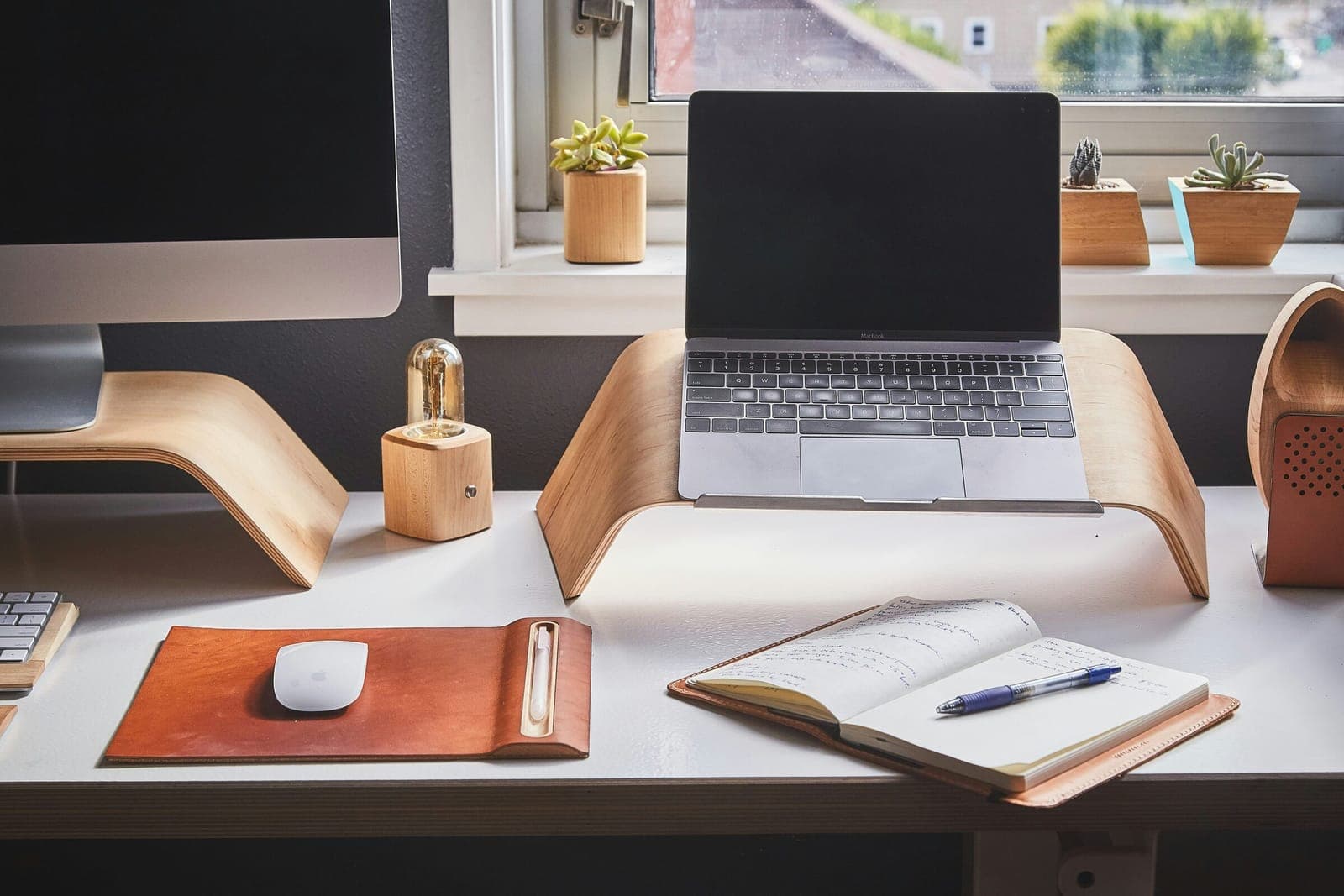 Bureau blanc devant une fenêtre, avec ordinateur portable, rehausseurs en bois et carnet ouvert