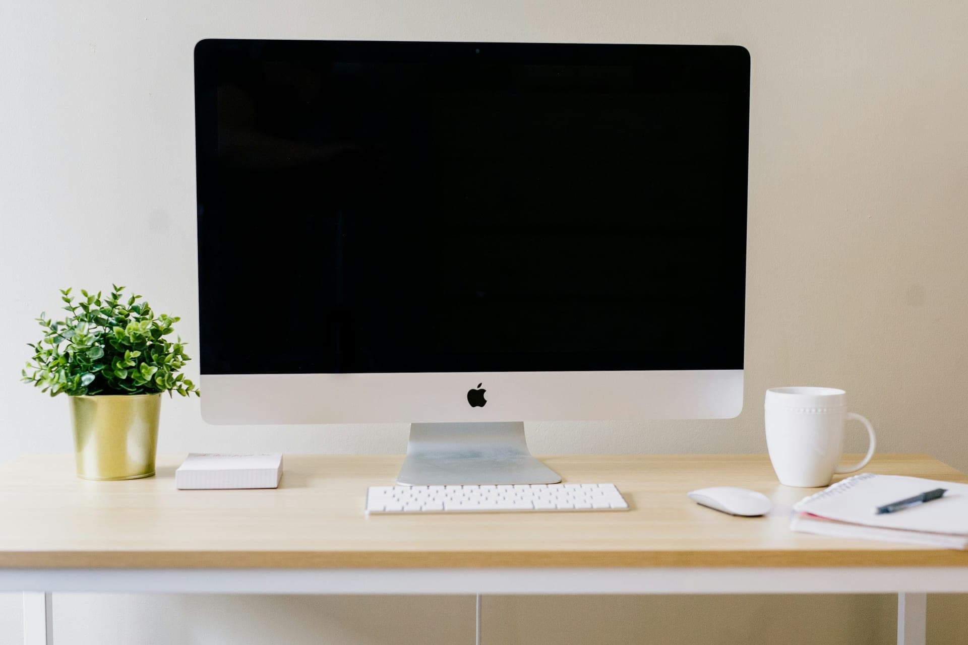 Dark screen, white keyboard and mouse on a pale wooden desk beside a plant and notebook