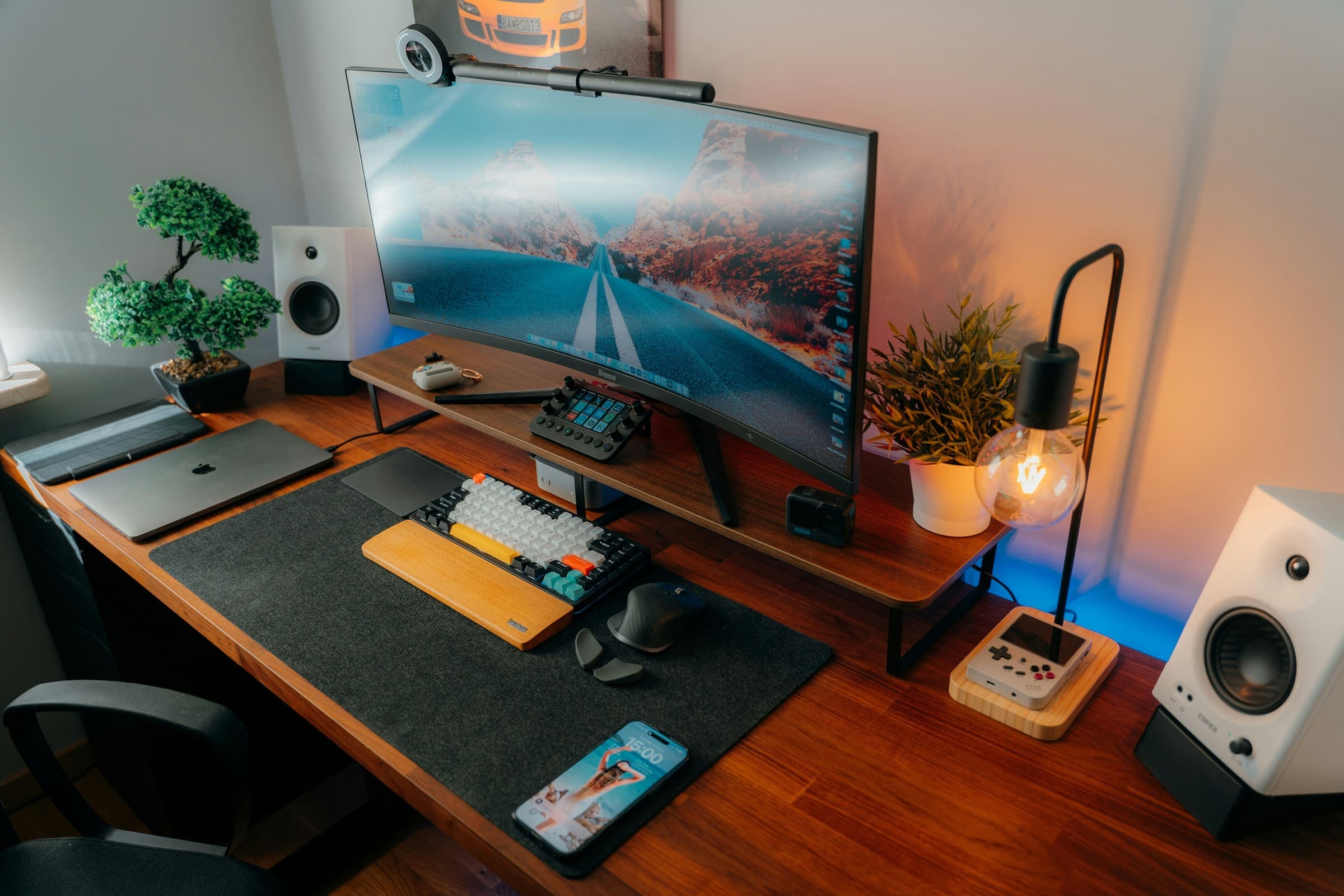 Wooden desk with a large felt desk mat, keyboard, mouse, wide monitor, lamp, speakers, and plant