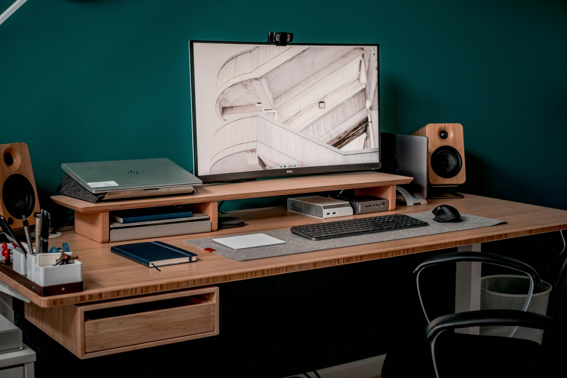 Bureau en bois avec étagère, écran, clavier et sous-main en feutre devant un mur vert profond.